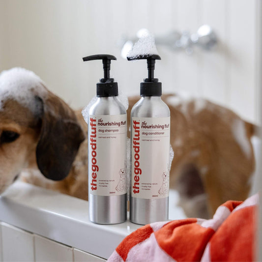 Two bottles of 'the good fluff' natural ingredients dog shampoo and conditioner on side of a bath with a dog in the background with soap bubbles