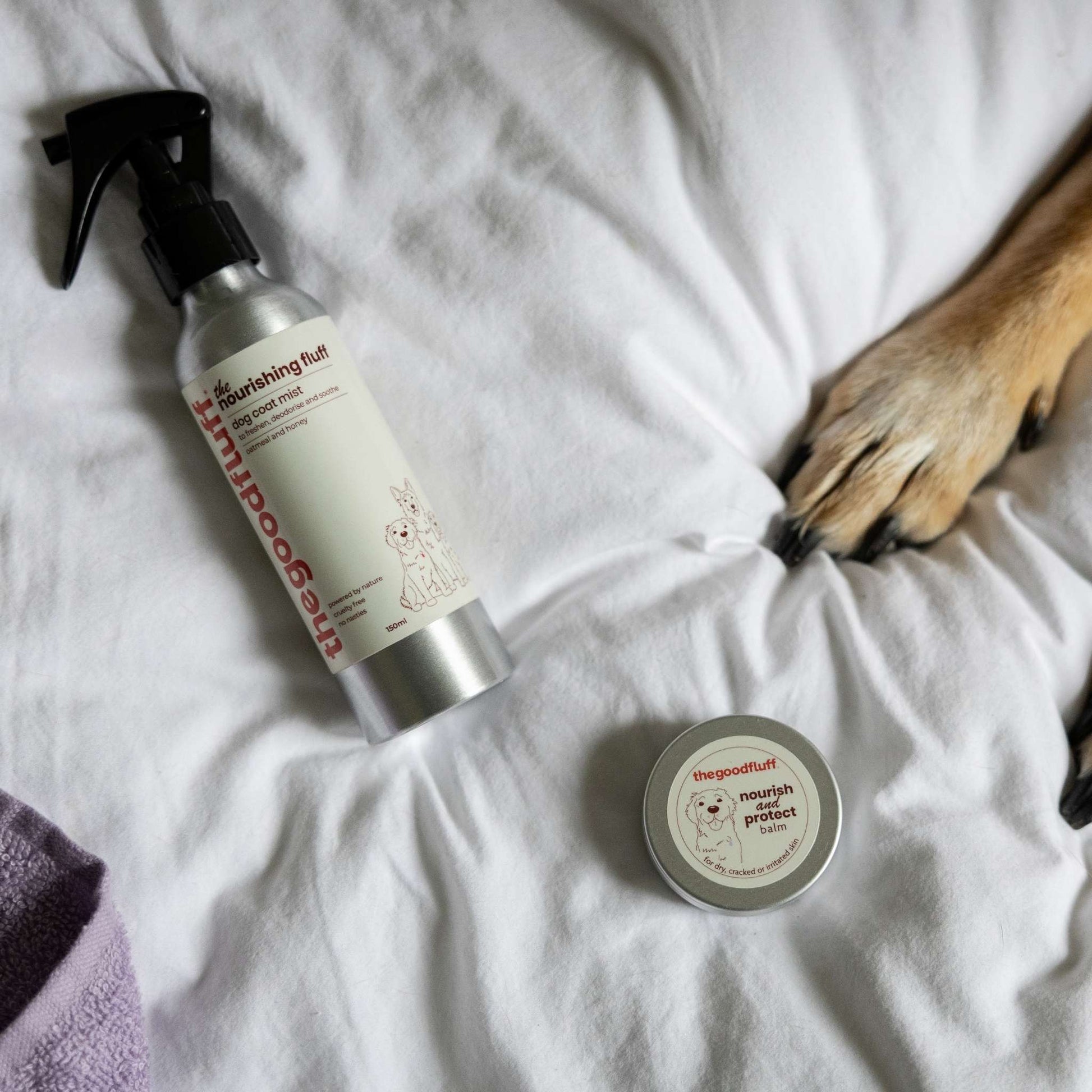 The Good Fluff Natural dog grooming products including a spray bottle and a paw balm tin on a white duvet with a dog's paw.
