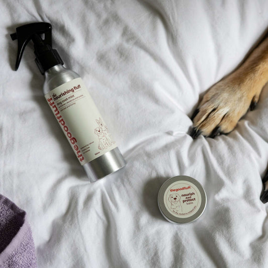 The Good Fluff Natural dog grooming products including a spray bottle and a paw balm tin on a white duvet with a dog's paw.