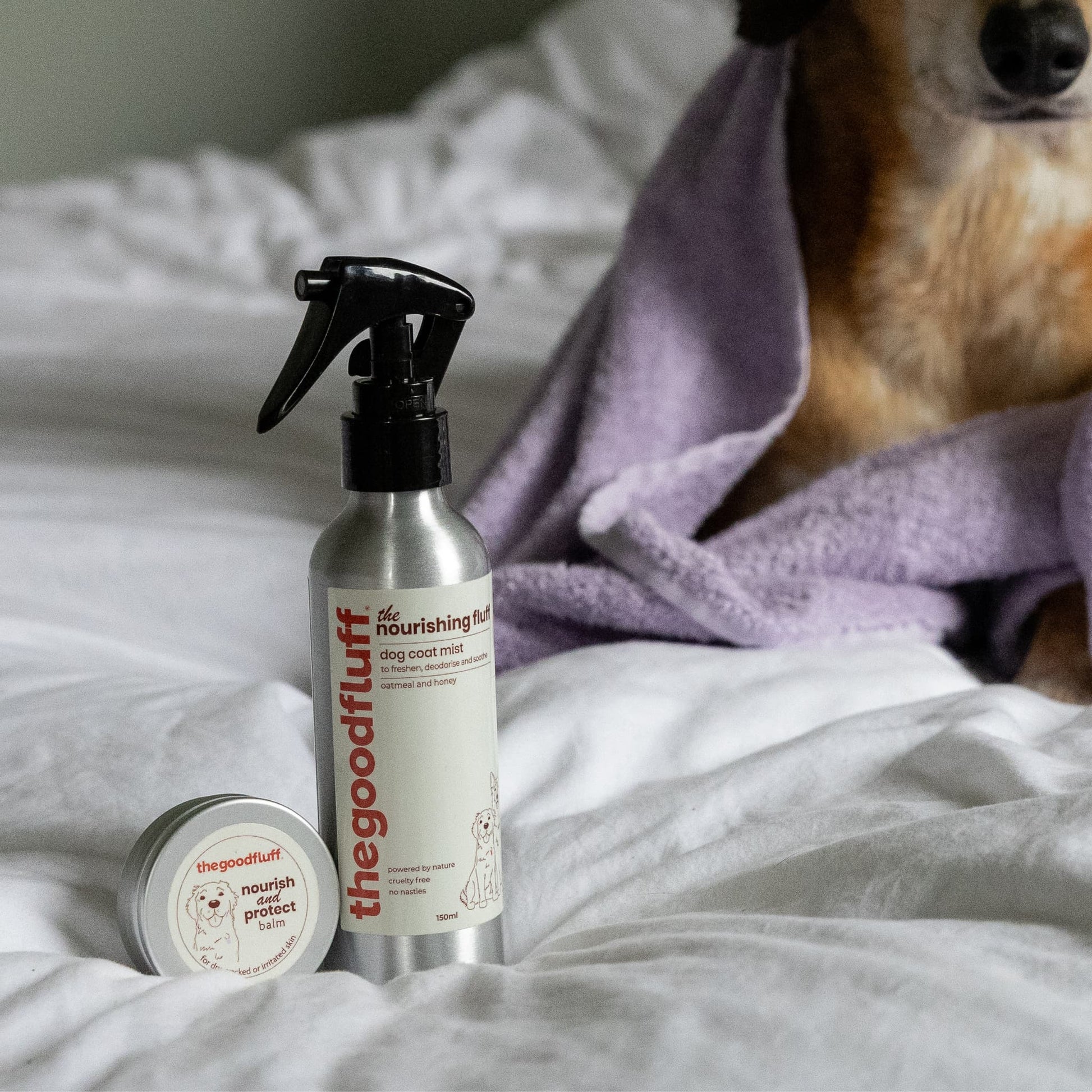 Dog sitting on a bed next to a bottle of 'the good fluff' natural dog coat mist and paw balm from the nourishing fluff collection