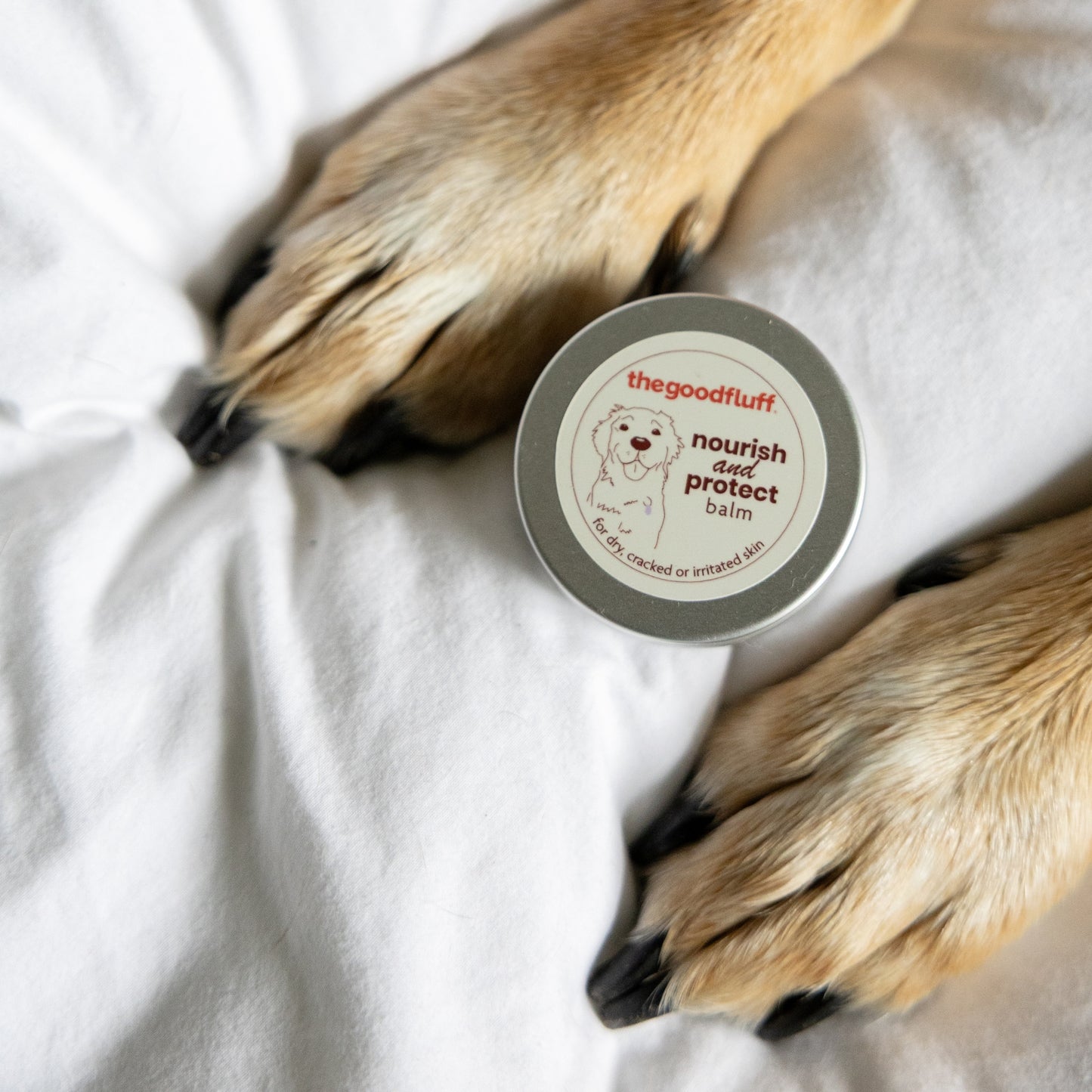 Balm tin of 'the good fluff' nourish and protect natural balm in between a dogs paws on a white duvet