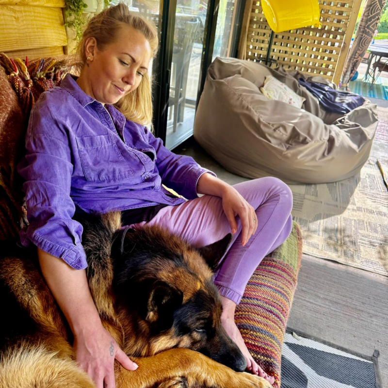 Kate the Co-founder of the good fluff in a purple shirt and pants sitting on a colourful chair with a German Shepherd cuddling